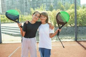 Kids and sports concept. Portrait of smiling girls posing outdoor on padel court with rackets and tennis balls photo