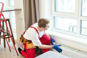 Full length shot of two professional plumbers, workers in uniform fixing or installing heating radiator. Construction, maintenance and repair concept. photo