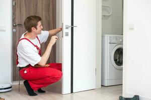 Repair Service Concept. Portrait of smiling handsome young locksmith in blue uniform installing door knob on front wooden entrance door at home or office workplace. photo