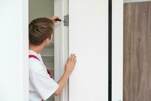 Young handyman installing a white door with an electric hand drill in a room photo