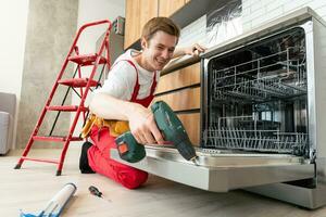 técnico o trabajador en uniforme instala lavavajillas dentro el cocina mueble. reparador vestir trabajador traje reparando mantenimiento de lavavajillas. Maestro en protector guantes reparar lavavajillas. foto