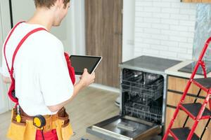 Technician or worker in uniform installs dishwasher into the kitchen furniture. Repairman wear worker suit repairing maintenance of dishwasher. Master in protective gloves fix dishwasher. photo