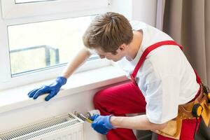 Young handsome professional plumber worker installing heating radiator in an empty room of a newly built apartment or house. Construction, maintenance and repair concept photo