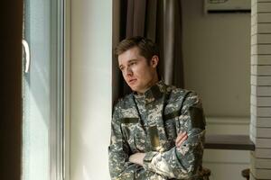 Depressed and sad soldier in green uniform with trauma after war standing near the window photo