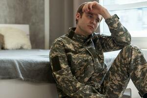 estresado veterano soldado absorto en depresivo pensamientos y recuerdos. triste Deprimido joven militar hombre en camuflaje uniforme sentado en sofá y pensando acerca de guerra. ptsd y terapia concepto. foto