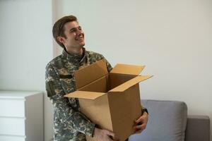 Smiling man in camouflage holding cardboard box in new apartments photo