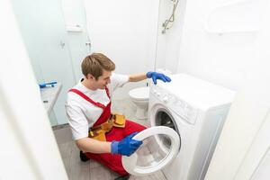 The young handsome repairman in worker suit with the professional tools box is fixing the washing machine in the bathroom. photo