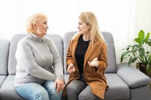 Beautiful senior mom and her adult daughter are hugging and smiling while sitting on couch at home photo