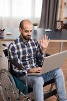 Disabled freelancer in wheelchair during a video call working from home. Businessman waving during video conference, working remotely from home on laptop. photo