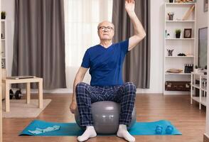 Elderly man training at home, doing arm exercises. Old person pensioner online internet exercise training at home sport activity with dumbbell, resistance band, swiss ball at elderly retirement age. photo