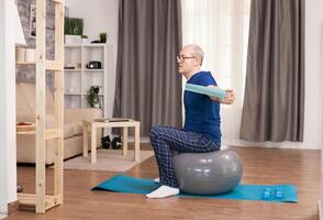 Pensioner doing sport with resistance band sitting on swiss ball. Old person pensioner online internet exercise training at home sport activity with dumbbell, resistance band, swiss ball at elderly retirement age. photo