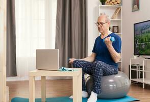 Senior man training with online coach sitting on swiss ball in the middle of the room. Old person pensioner online internet exercise training at home sport activity with dumbbell, resistance band, swiss ball at elderly retirement age. photo