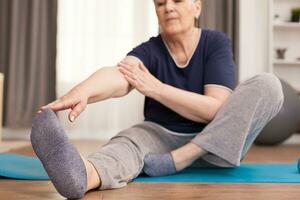 Modern grandmother doing stretching at home oon yoga mat. Old person pensioner online internet exercise training at home sport activity with dumbbell, resistance band, swiss ball at elderly retirement age photo