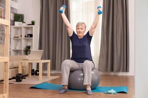 Grandmother with healthy lifestyle practicing fitness at home. Old person pensioner online internet exercise training at home sport activity with dumbbell, resistance band, swiss ball at elderly retirement age photo