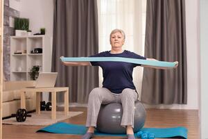 Ambitious woman doing sports at home with resistance band. Old person pensioner online internet exercise training at home sport activity with dumbbell, resistance band, swiss ball at elderly retirement age photo