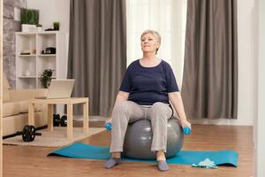 Senior woman doing exercise for arms with dumbbells in apartment. Old person pensioner online internet exercise training at home sport activity with dumbbell, resistance band, swiss ball at elderly retirement age photo