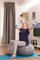 Senior woman using balance ball and dumbbells sitting on yoga mat at home. Old person pensioner online internet exercise training at home sport activity with resistance band, swiss ball at elderly retirement age photo