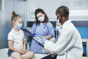 Nurse in blue scrubs filling out a clipboard with information on the girl. Face masks were worn by the patient, her mother, and the multicultural medical personnel. An atmosphere of professionalism. photo