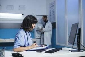 Professional medic looking at computer screen and medical files for healthcare. Nurse reviewing information on pc monitor and documents for assistance and support, doing overtime work. photo