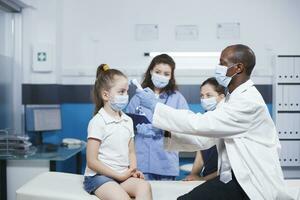 africano americano médico vistiendo un máscara y laboratorio Saco cheques el temperatura de un joven muchacha. masculino pediatra usos un termómetro y consulta con el madre de el paciente. foto