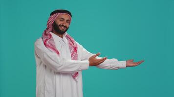 Muslim adult in traditional gown and ghutra presenting sides with hands and posing for studio shot. Arabic joyful man promoting something while showing marketing advertisement aside. video