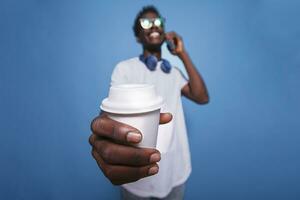 Joyful black man holds a coffee cup close to the camera. Casual african americam adult on a phone call, wearing sunglasses, and holding a beverage over an isolated background. photo