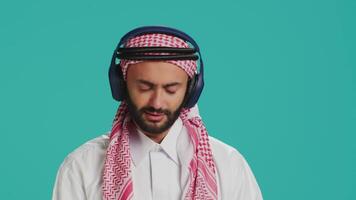 Groovy man dancing on camera, listening to modern tunes on headset and having fun in studio. Middle eastern man in traditional attire showing funky dance moves and singing songs. video