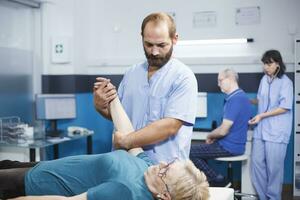 mayor paciente consulta especialista asistente, recepción médico ayuda para músculo dolor. enfermero facultativo realiza examen, estiramientos brazos, y proporciona fisioterapia. foto