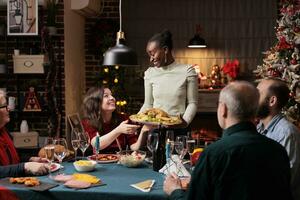 personas disfrutando Navidad víspera cena a hogar con festivo decoraciones y Navidad árbol cerca chimenea. familia y amigos celebrando invierno fiesta temporada con tradicional alimento. foto