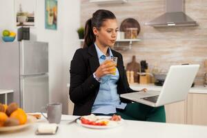 Business woman taking a break and relaxing watching a video clip, drinking orange juice. Young freelancer in the kitchen talking on a video call with her colleagues from the office, using modern internet technology photo