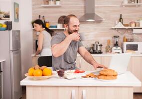 Happy entrepreneur working from home and talking on webcam with colleagues. Eating breakfast in kitchen, wearing pajamas enjoying roasted bread with butter. Freelancer working online via internet using modern digital technology photo