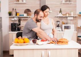 contento Pareja surf en Internet utilizando ordenador portátil durante desayuno en cocina. casado marido y esposa en pijama utilizando Internet web en línea moderno tecnología, sonriente y contento en el Mañana. leyendo Noticias hojeada Internet foto