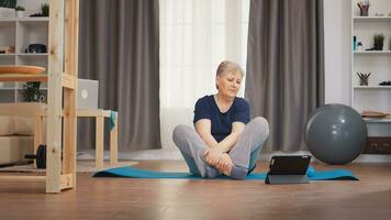Senior woman stretching body watching online lesson sitting on yoga mat. Online learning and study, active healthy lifestyle sporty old person training workout home wellness and indoor exercising photo