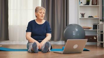 Old woman sitting on yoga mat stretching body watching exercise lesson on laptop. Retired old person learning new tehnology lessons online using laptop, healthy lifestyle combined with sport photo