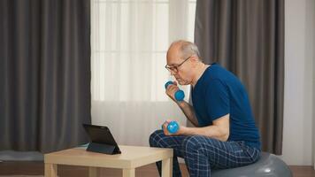 Elderly aged male exercising with dumbbells sitting on swiss ball during online training. Old person pensioner healthy training healthcare sport at home, exercising fitness activity at elderly age photo