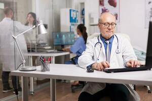 Doctor with grey hair in clinic cabinet working on computer and young medic discussing with woman patient on hospital corridor. photo