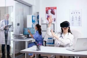 retrato de profesional mujer médico en vr lentes sentado en privado hospital gabinete y haciendo gestos utilizando virtual realidad innovaciones mientras equipo de doctores trabajando en antecedentes foto