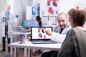 Young doctor explaining heart disease pointing on laptop sitting in hospital room talking with senior woman. Team of doctors working in background using clipboard, body scan and xray of sick patient photo