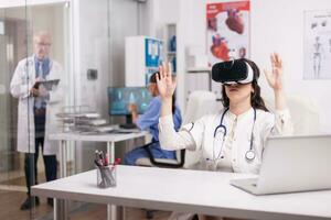 Young doctor using augmented reality goggles in hospital office while senior medic is taking notes on clipboard in clinic corridor and nurse working on computer. photo
