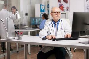 Senior doctor with stethoscope using pc in hospital cabinet while young medic wearing white coat checking report with patient on hospital corridor and nurse in blue uniform. photo