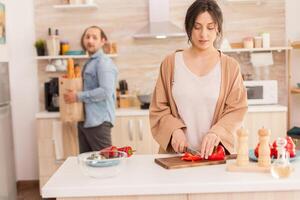 Wife cutting salad ingredient while husband is holding grocery bag with fresh vegetables from market. Funny happy in love couple at home spending time together, cooking healthy and smiling photo