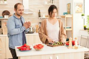 Woman cutting cucumbers for healthy salad in kitchen while having a conversation with husband. Happy in love cheerful and carefree couple helping each other to prepare meal photo