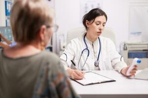 Medic writing senior patient diagnosis after measuring body temperature with thermometer gun in hospital office. Doctor with stethoscope wearing white coat. photo