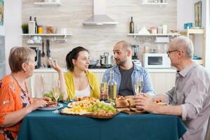 Multi generation family eating meal around the table in kitchen. Senior man holding wine glass. Tasty seasoned potatoes. photo