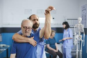 Medical nurse stretching muscles and treating physical injuries by elevating arm of elderly person. At physiotherapy consultation, male osteopath does mobility exercises with senior patient. photo