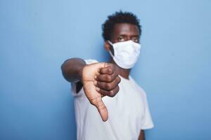 African american adult doing thumbs down gesture with right hand in studio. Black person showing sign while staring at camera and wearing face mask to protect from coronavirus pandemic. photo