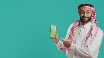 Middle eastern man points at greenscreen display on smartphone, showcasing isolated copyspace template on mobile device screen. Young person in islamic costume shows blank chromakey. video