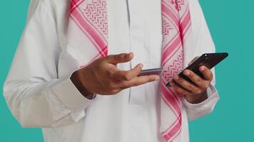 muslim man tillsats kort information på webbsida, håller på med uppkopplad handla session medan han bär traditionell islamic klädsel med huvudduk. person betalande för objekt på webb Lagra, bank service video