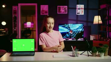 Cheerful child in home studio presenting advertisement from sponsoring brand on green screen laptop. Gen Z girl using chroma key device to present promotion from media partner to followers video