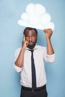 Professional businessman with hand on forehead is holding white paper idea cloud and standing in front of isolated blue background. African american person brainstorming under thought bubble. photo